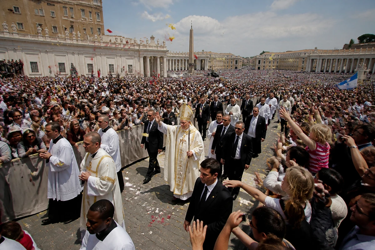 Processione con un pontefice in abiti bianchi tra due ali di folla in una grande piazza, con petali in aria