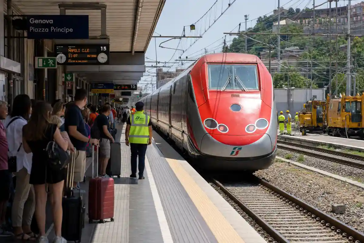 Treno ad alta velocità rosso in stazione a Genova Piazza Principe, passeggeri in attesa e operai al lavoro sui binari